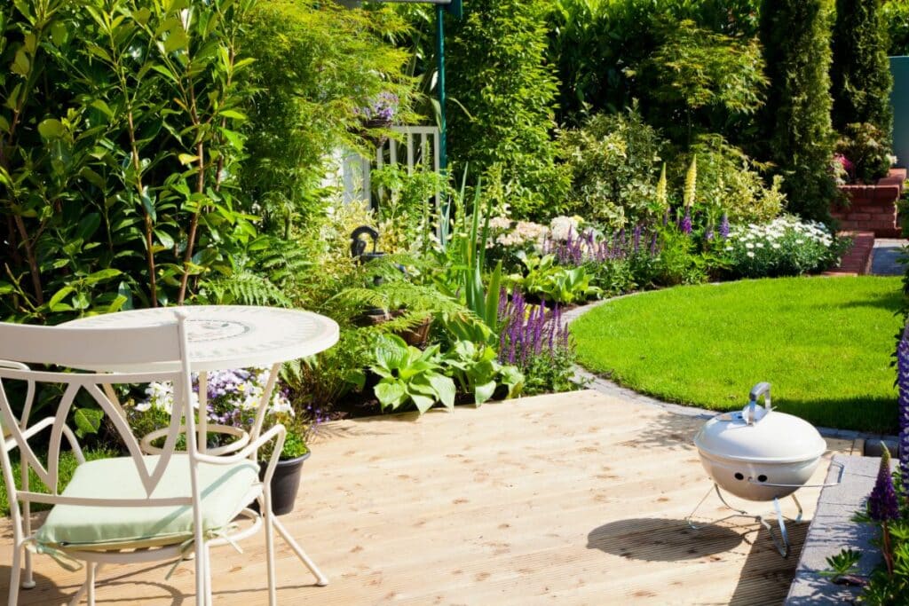 A small patio with a white metal table and chair overlooks a lush garden filled with green plants, colourful flowers, manicured grass, and a white barbecue on the wooden decking.