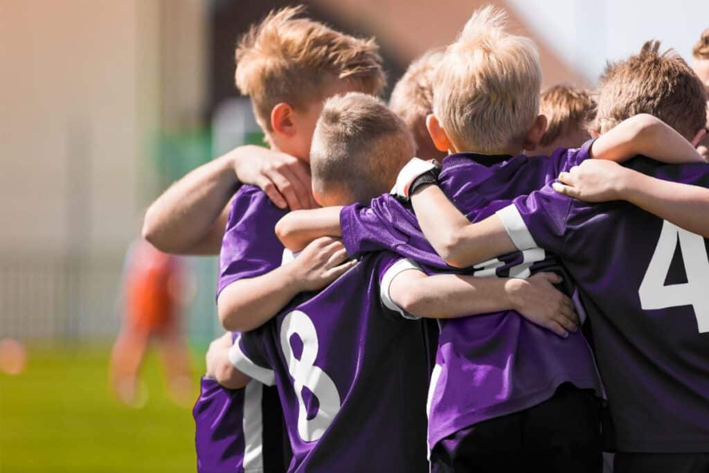A group of young boys wearing purple football jerseys huddle together with their arms round each other on a grassy pitch, showing the spirit of a true team player and unity before a match.