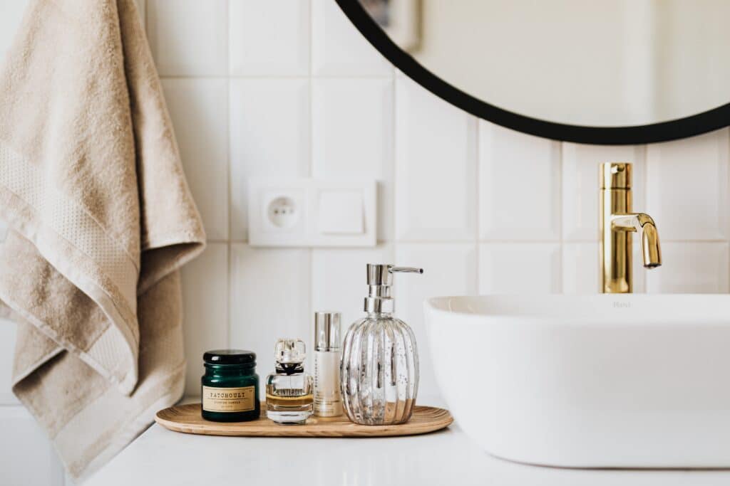 A beautiful bathroom countertop with a white sink, gold tap, and a tray holding toiletries. Beige towels hang on the wall, and a round mirror is partially visible above the white tiled walls.