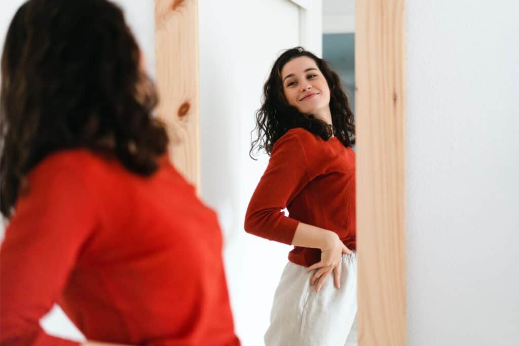 A woman with dark, curly hair wearing a red top and light trousers smiles confidently at herself in a full-length mirror—Boost Your Confidence with Shapewear.