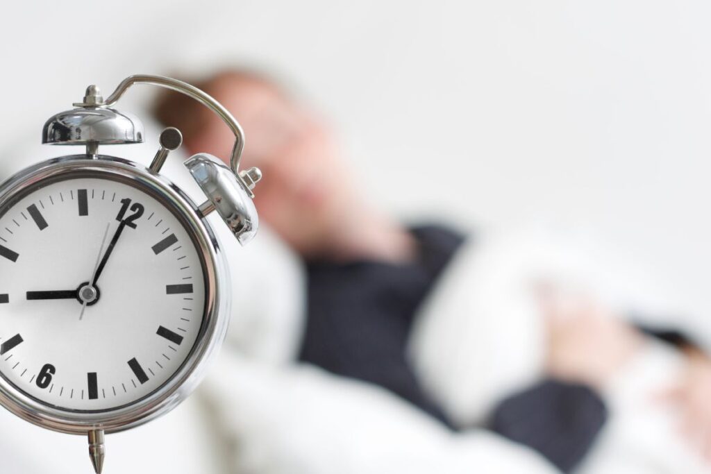 A close-up of a classic alarm clock showing 7:00, with a blurred teenager lying in bed in the background. The scene suggests waking up or getting ready to start the day.