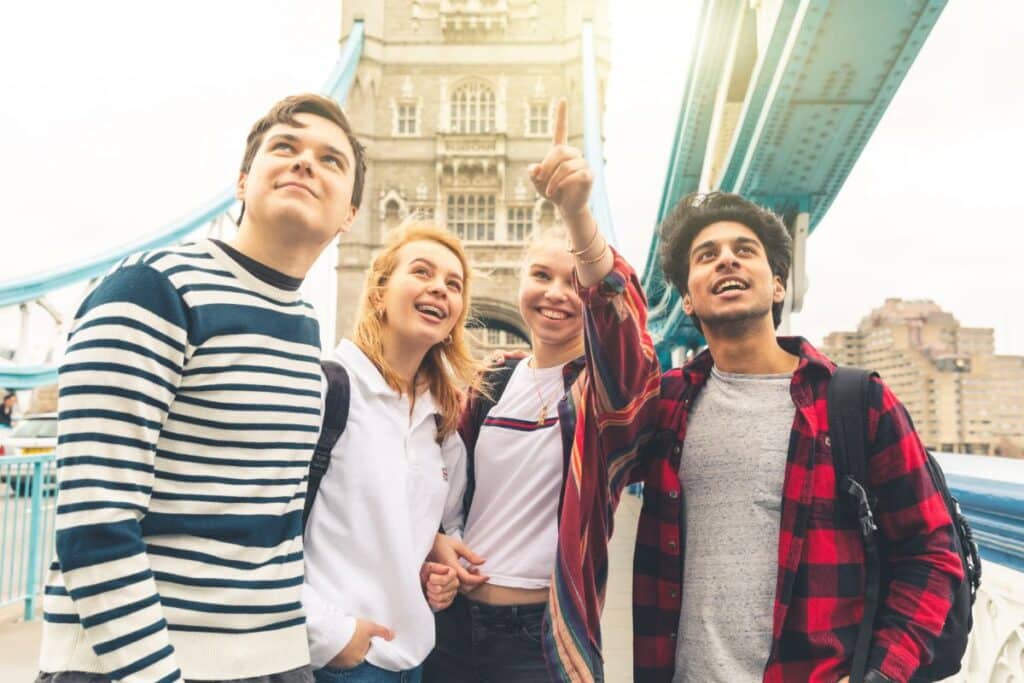 Four young adults stand on a bridge, smiling and looking up excitedly as they discuss planning a school trip. One person points at something in the distance with Tower Bridge’s iconic towers and blue railings in the background.