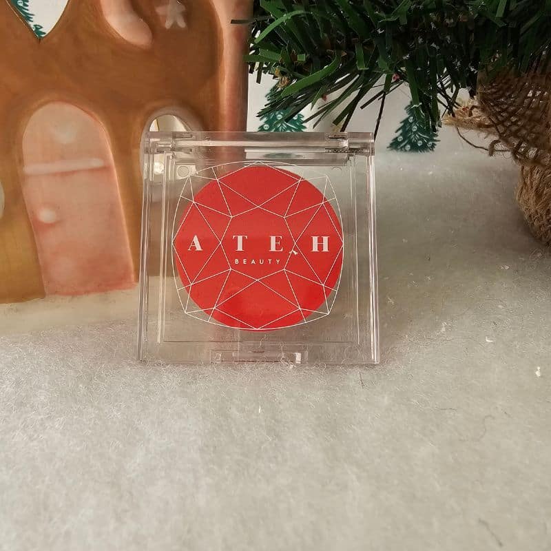 A red geometric-patterned compact labelled "ATEH BEAUTY" rests on a snowy surface near a ceramic house and pine tree branches, evoking a festive Christmas atmosphere.