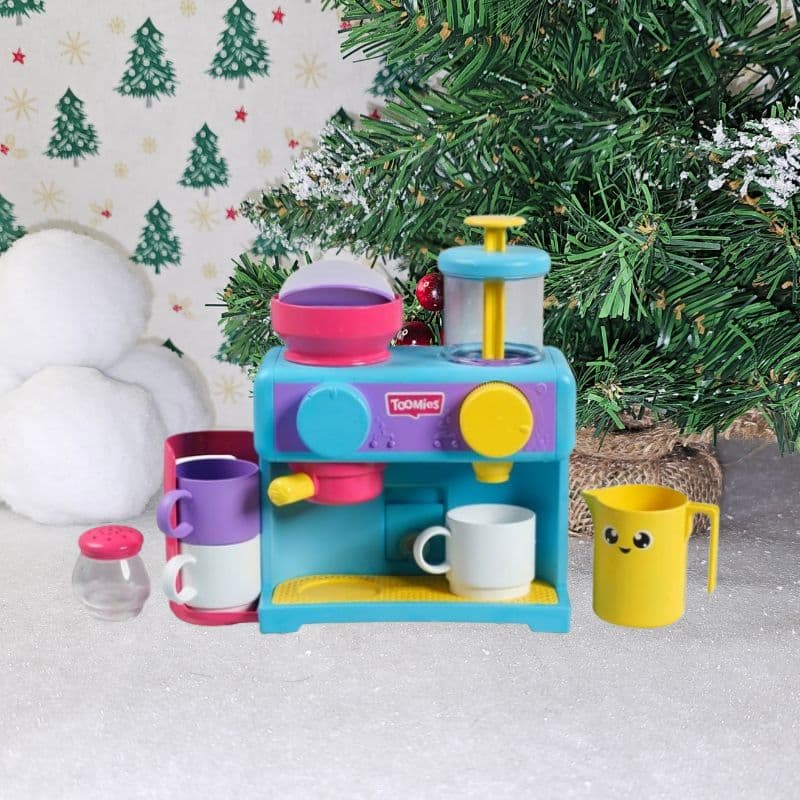 A colourful toy coffee machine with cups and accessories sits on a snowy surface, in front of a decorated Christmas tree and snowballs, capturing the joyful spirit of Christmas with festive green tree wallpaper in the background.