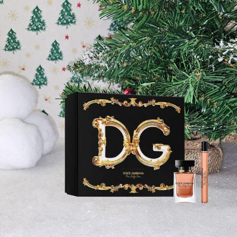 A Dolce & Gabbana gift set with a black and gold box, perfume bottle, and make-up item is displayed in front of a decorated Christmas tree and festive background, perfect for celebrating the magic of Christmas.