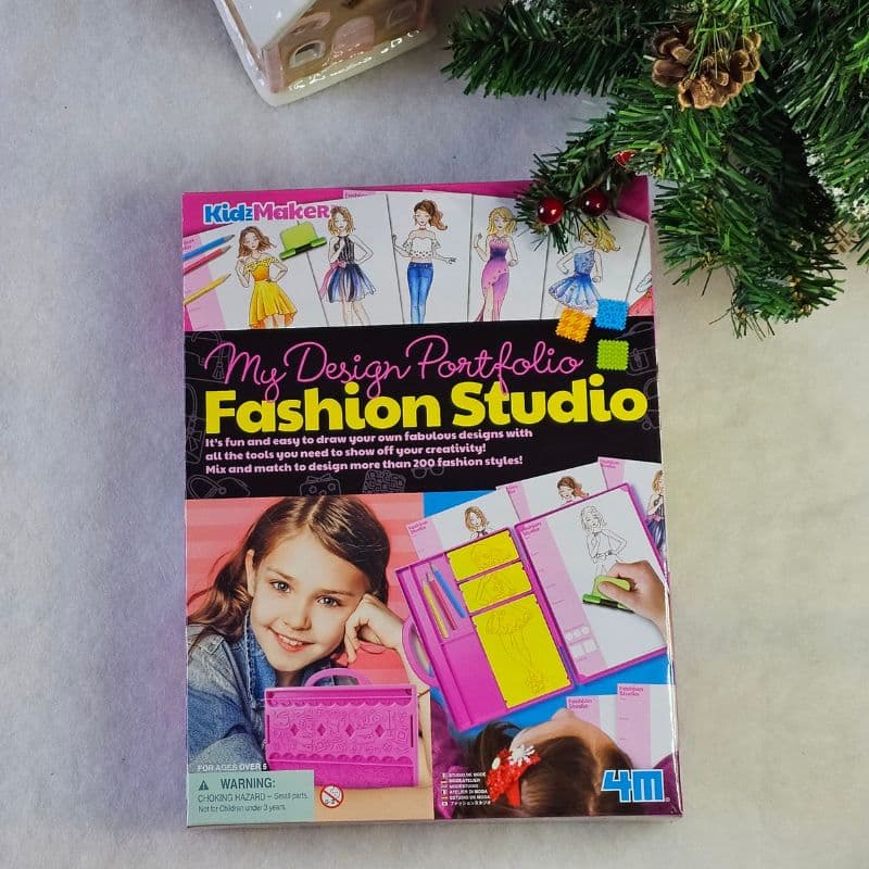 A box of "My Design Portfolio Fashion Studio" craft kit on a white surface sits beside a small pine branch with pine cones and a gold bauble, creating a festive Christmas vibe. The kit features fashion sketches and a smiling girl on the cover.