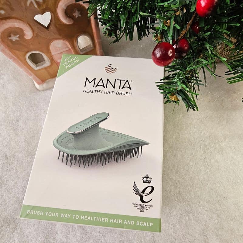 A MANTA Healthy Hair Brush in its packaging sits on a white surface next to a decorated Christmas tree and a gingerbread house-shaped ornament, creating a festive scene. The packaging highlights flexible bristles for gentle, healthy hair and scalp care.