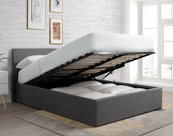 A grey upholstered ottoman bed with its mattress lifted to reveal spacious storage space underneath, set in a modern bedroom with light-coloured walls, framed art, and a large window.