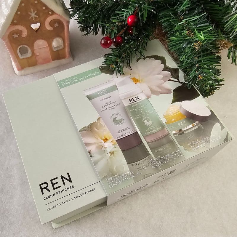 A REN Clean Skincare Christmas gift set is placed on a white surface next to a decorated Christmas tree and a gingerbread house candle holder. The box displays images of skincare products and festive floral graphics.