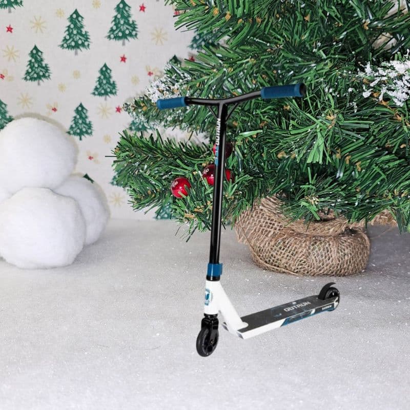 A small scooter sits on white snow-like ground next to a decorated Christmas tree, bringing a festive Christmas feel, with snowballs and a wall covered in green tree patterns in the background.