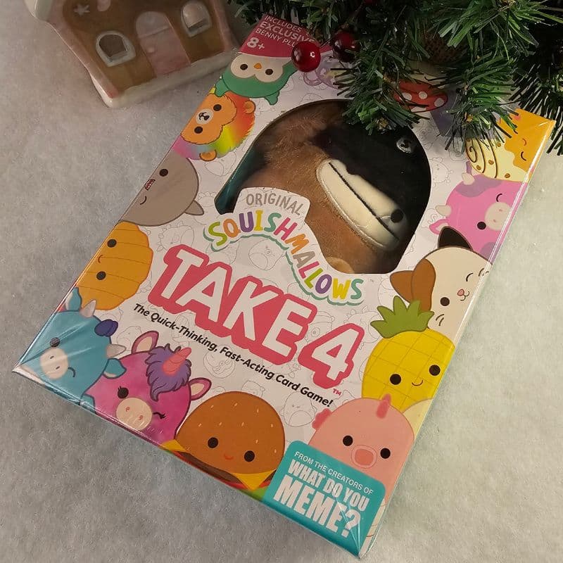 A boxed Squishmallows-themed card game called "Take 4" sits beneath a Christmas tree branch on a white surface, bringing festive Christmas fun with colourful Squishmallow characters illustrated on the packaging.