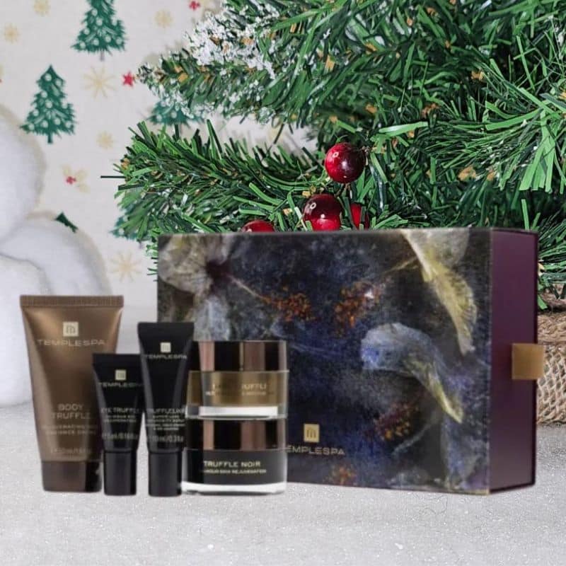 A set of TEMPLESPA skincare products is displayed in front of a decorated Christmas tree, with a festive Christmas gift box and small tubes and jars arranged neatly on a white surface.