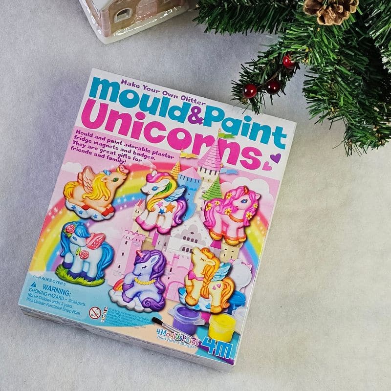 A box labelled "Mould & Paint Unicorns" sits on a grey surface beside a Christmas tree and a small pink house. The colourful box highlights fun unicorn crafts and castles, making it a festive arts and crafts gift for children this Christmas.