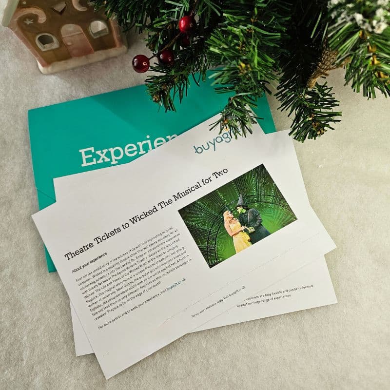 Two theatre ticket vouchers for "Wicked The Musical" rest on a teal "Experience" envelope beside a festively decorated Christmas tree branch and a small gingerbread house, creating a perfect holiday gift scene.