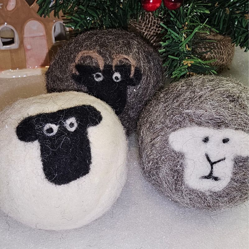 Three felted wool balls with sheep faces, in white, brown, and grey, are displayed in front of a small artificial Christmas tree and a wooden house decoration for a festive Christmas touch.