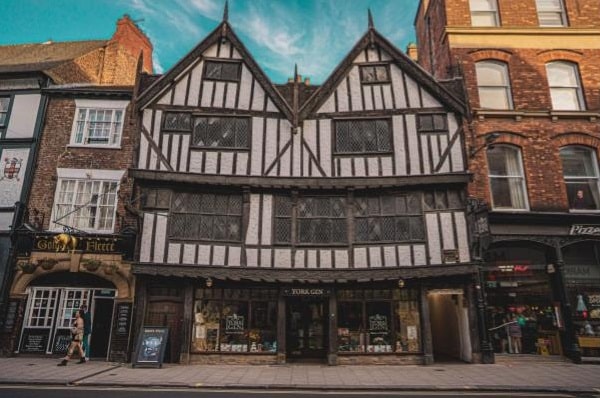 A historic, black-and-white timber-framed Tudor-style building with steep gabled roofs and lattice windows, ideal for those buying a home in Yorkshire, stands between brick structures on a city street with shops under a blue sky.