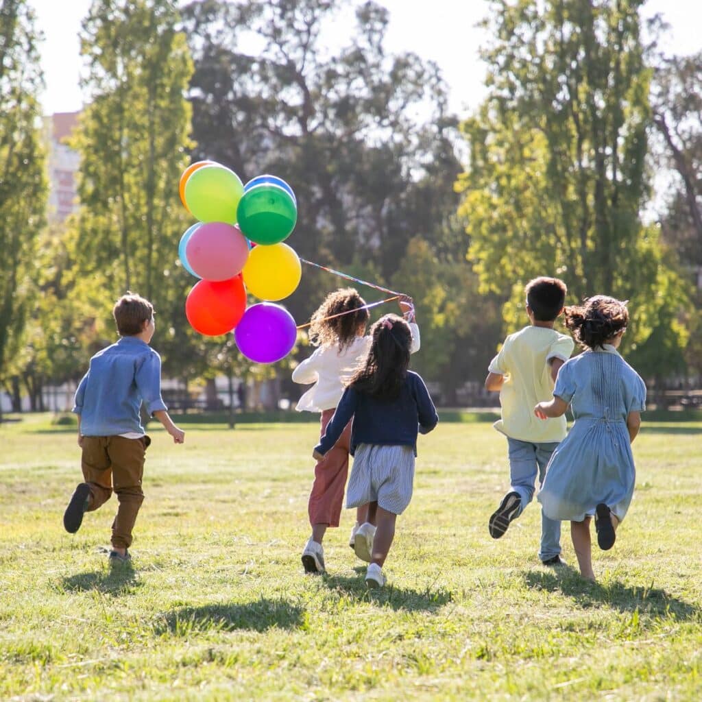Five children run across a grassy field, holding colourful balloons. The bright, sunny scene with tall trees in the background highlights the joy and freedom that nurture healthy childhood development outdoors.