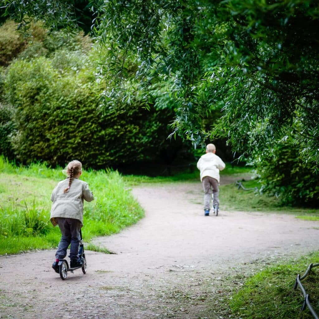 Two children ride scooters along a winding dirt path surrounded by lush green trees and bushes in a park. The kids, dressed in light jackets and trousers, enjoy the peaceful, natural setting as they zip along on their scooters.