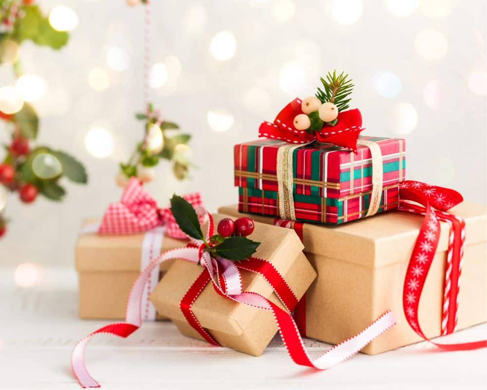 Three Christmas gift boxes, two in plain brown paper with red and white ribbons, and one in red tartan wrapping with a festive decoration, are arranged on a white surface with a blurred, sparkling background.