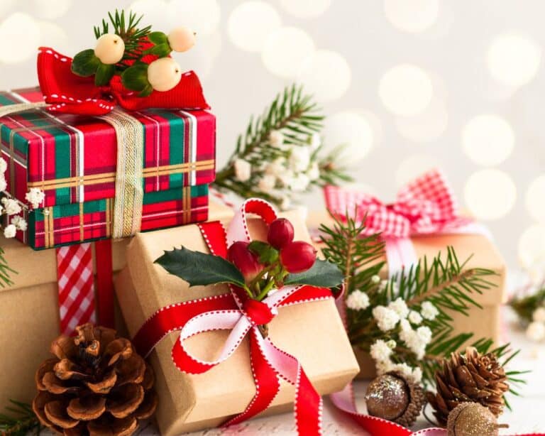 Three Christmas presents wrapped in tartan and brown paper with festive ribbons, holly, and pine decorations sit surrounded by pine cones and greenery, creating a charming Christmas scene against a soft, glowing background.