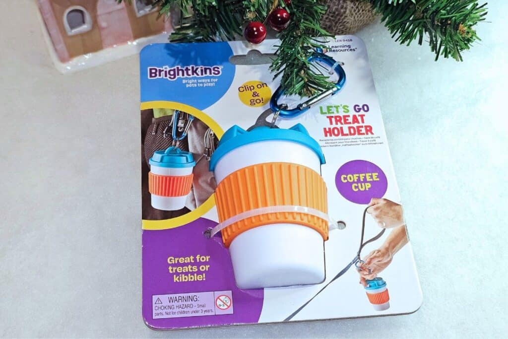 A Brightkins treat holder for dogs shaped like a coffee cup, attached to its packaging with a blue clip. The packaging shows a hand holding the holder and text describing it as great for treats or kibble. Some tree branches are visible above.