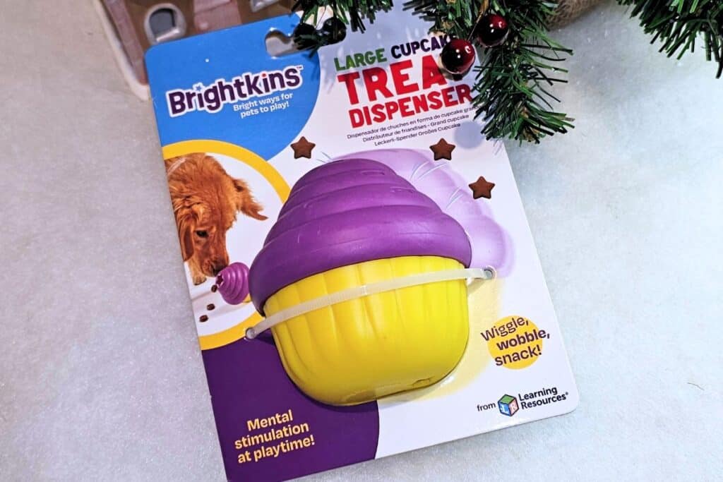 A Brightkins Large Cupcake Treat Dispenser for dogs in purple and yellow packaging, featuring a photo of a happy dog and product details. A green Christmas tree branch is visible in the top right corner.