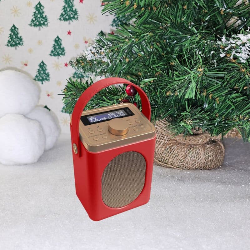A red and gold portable radio sits on a snowy surface beside a decorated Christmas tree and white baubles, with festive Christmas wallpaper featuring green Christmas trees in the background.