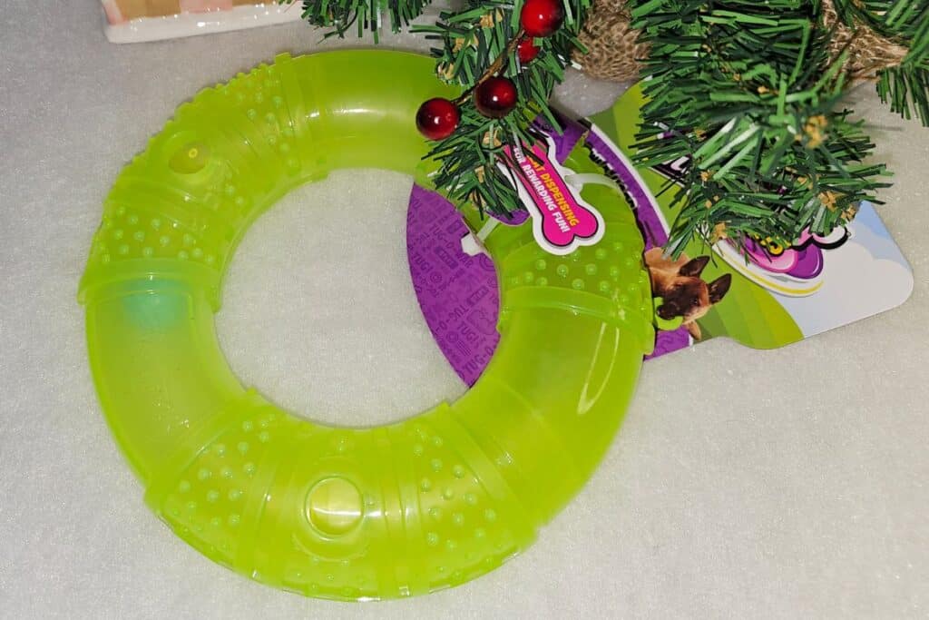 A neon green textured dog chew ring toy lies partially under a small decorated Christmas tree on a white surface. The packaging, featuring an image of dogs, is also visible.