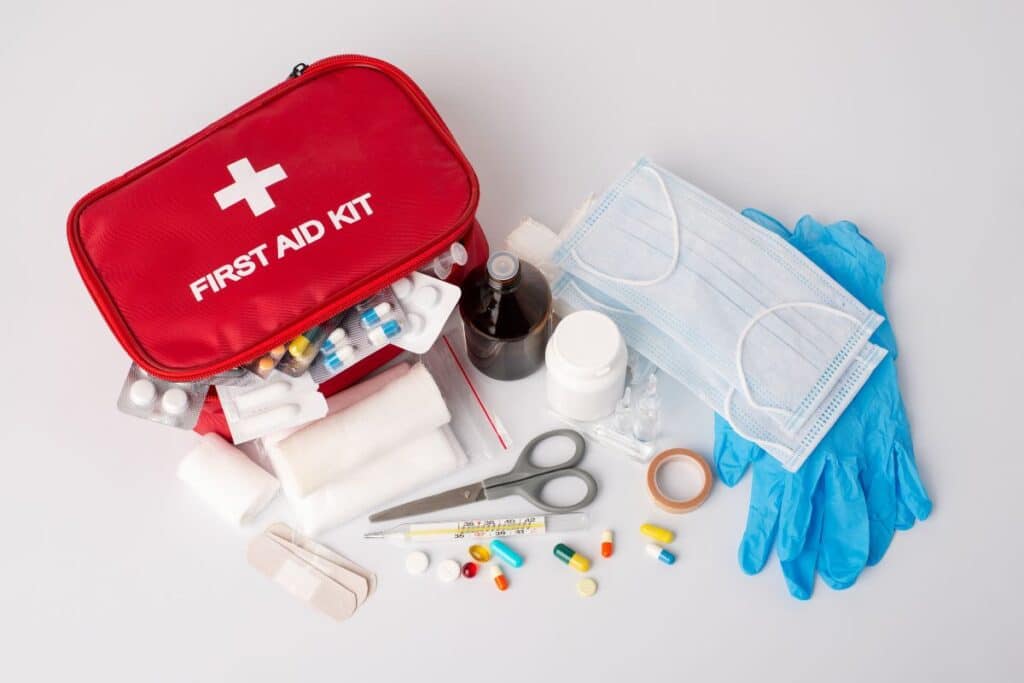 A red first aid kit surrounded by essential medical supplies, including tablets, bandages, scissors, tape, gloves, masks, medicine bottles, a thermometer, and blister packs on a white surface.