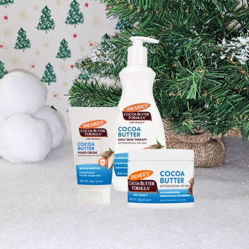 Three Palmer’s Cocoa Butter Formula skincare products are displayed in front of a decorated Christmas tree, with snowballs on the left and a snowy, festive Christmas background.