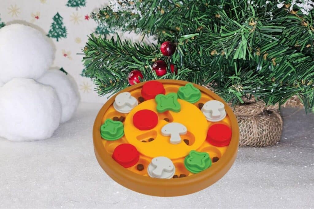 A toy pizza with plastic toppings sits on snowy ground beside a decorated Christmas tree and snowballs, whilst playful dogs frolic nearby, all set against a festive backdrop featuring green stars and trees.