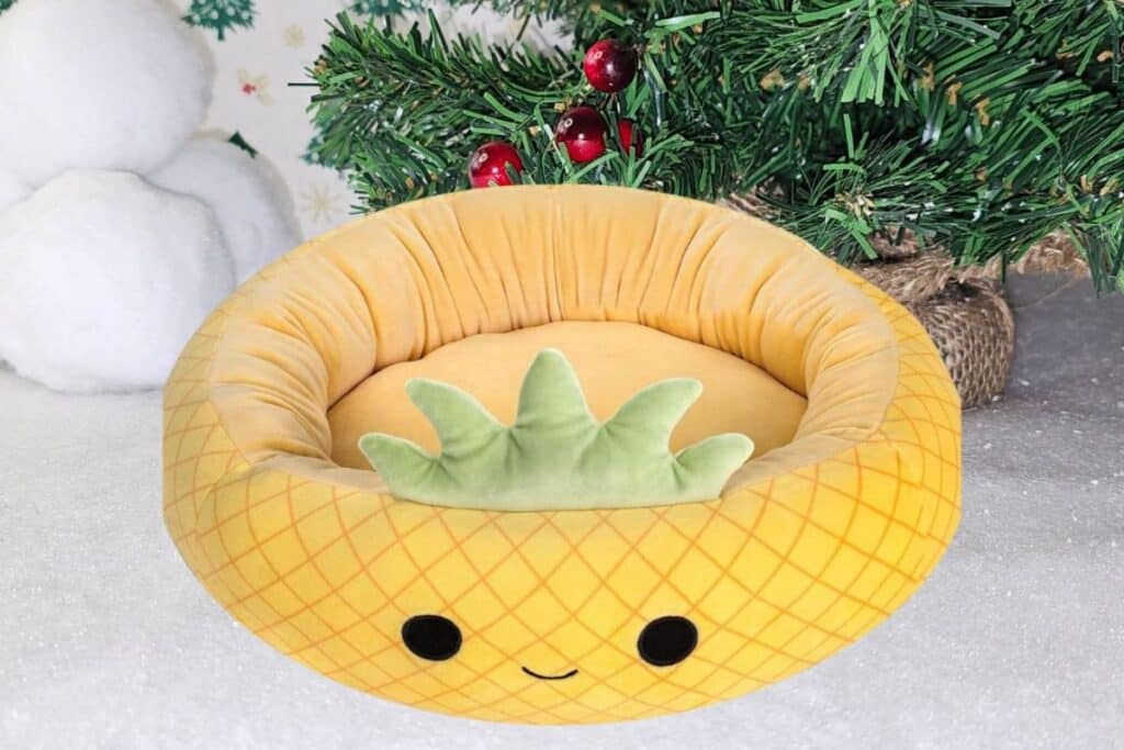 A round, yellow pet bed for dogs, designed to look like a smiling pineapple with a green leafy top, sits on a snowy surface beside a decorated Christmas tree.