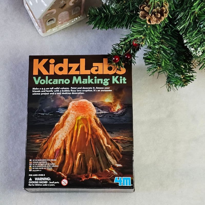 A KidzLabs Volcano Making Kit box sits on a table next to a decorated Christmas tree, making it a perfect Christmas present. The box features an erupting volcano and includes instructions and safety warnings on the front.