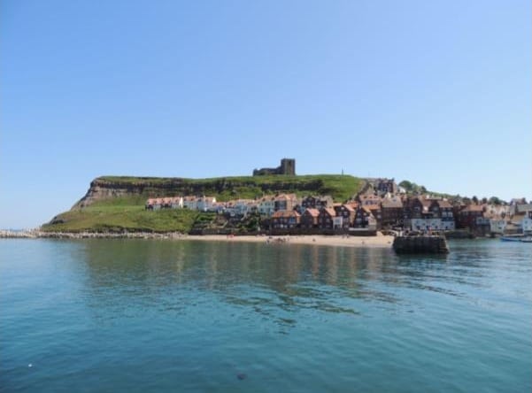 A coastal town with colourful houses along the shore, a grassy hill crowned by castle ruins, and calm blue water in the foreground under a clear sky—perfect for those considering buying a home in Yorkshire.
