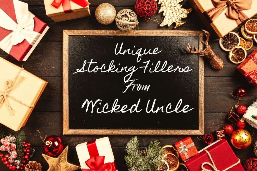 A blackboard with the words "Unique Stocking Fillers From Wicked Uncle" surrounded by stocking filler gifts, ornaments, and festive decorations on a dark wooden table.