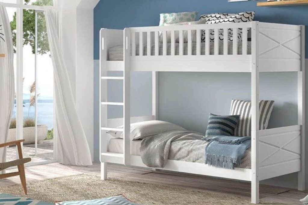 White wooden bunk beds with grey and blue bedding anchor this bright, coastal-themed bedroom. Sunlight pours through large windows overlooking water and trees, whilst a rug, pillows, and decor create a cosy retreat.