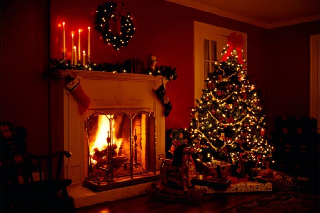 A cosy living room decorated for Christmas, featuring a lit fireplace with hanging stockings, glowing Christmas lights on a brilliantly adorned tree with baubles and presents beneath, and candles and a wreath on the mantelpiece.