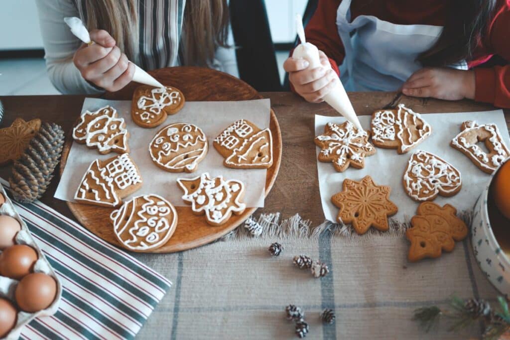 Two people decorate gingerbread biscuits with white icing at a wooden table. Surrounded by eggs, pine cones, and holiday-themed items, the scene captures the festive atmosphere of a birthday party in winter with intricately shaped biscuits.