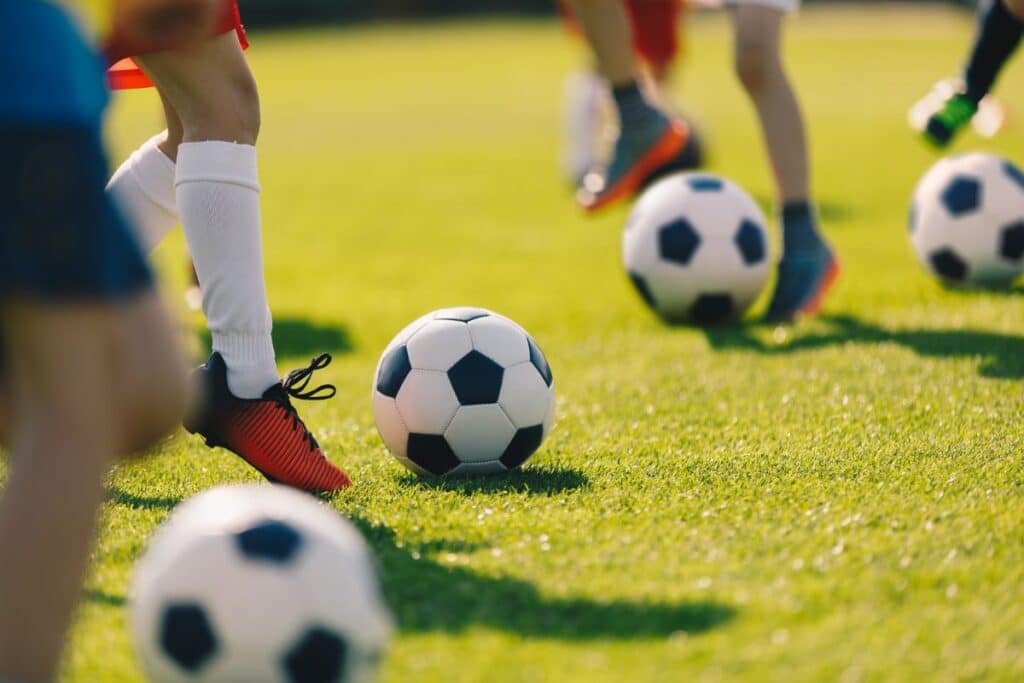 Children on a grassy field practising football lessons, each standing near a football. The focus is on their legs and feet in sports shoes as they get ready to kick the balls.