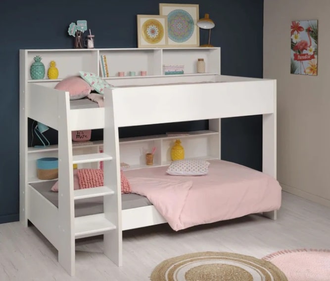 White loft bunk bed with built-in shelves adds style to a child's bedroom, featuring a grey lower bed and a pink upper bed. Decorative items, books, and pillows line the shelves; artwork and a lamp sit on the shelf above the bed.