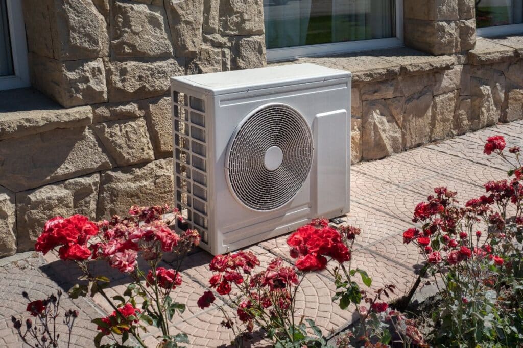 A white outdoor air conditioning unit sits on a patterned stone patio, surrounded by blooming red roses in front of a stone building wall—a perfect spot to consider the pros and cons of a heat pump for your home.
