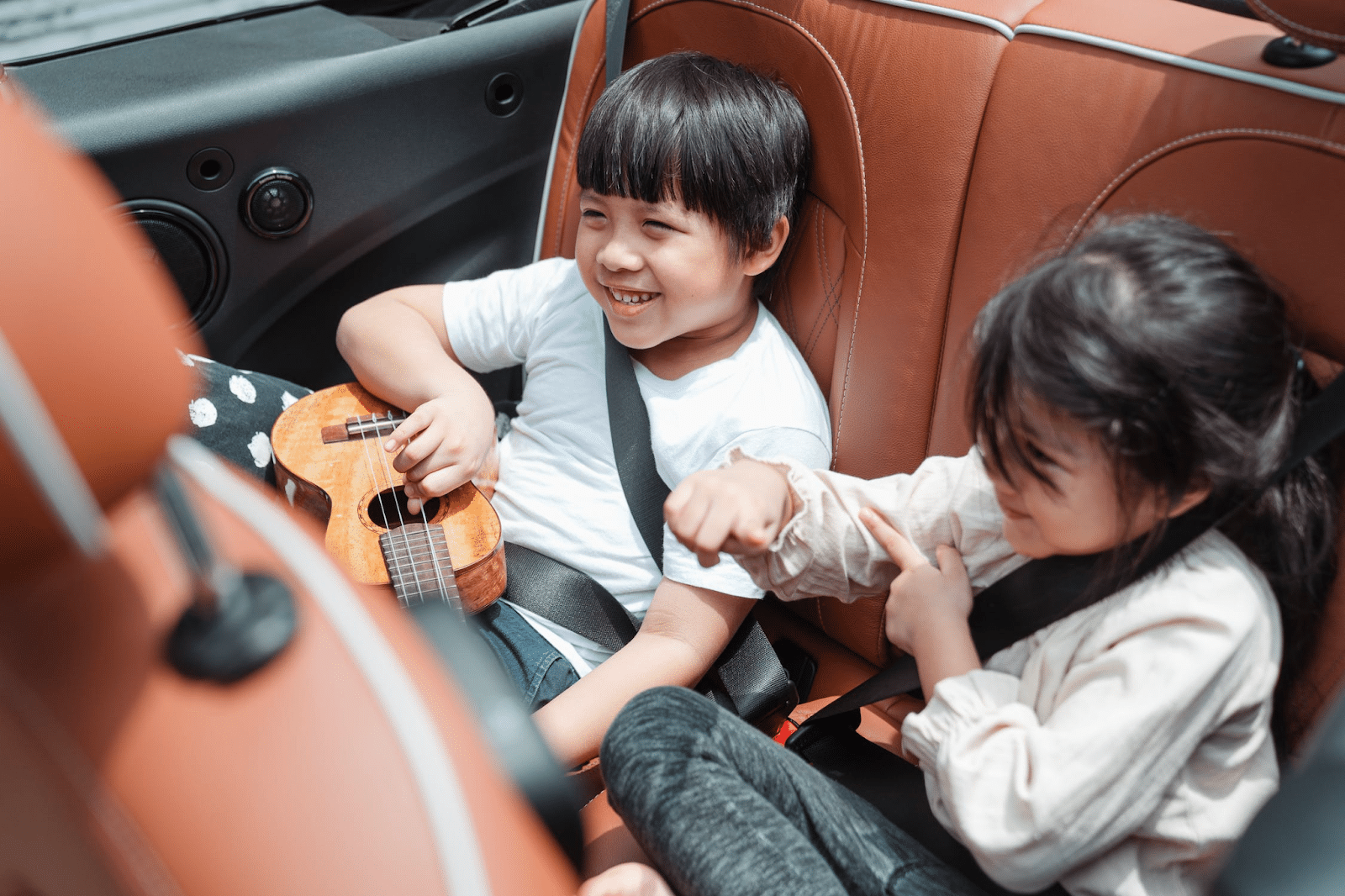 Two smiling children sit in the back seat of a car; the boy is holding a small ukulele while the girl playfully points at him. Both are wearing seat belts and look happy, enjoying a joyful family moment together.
