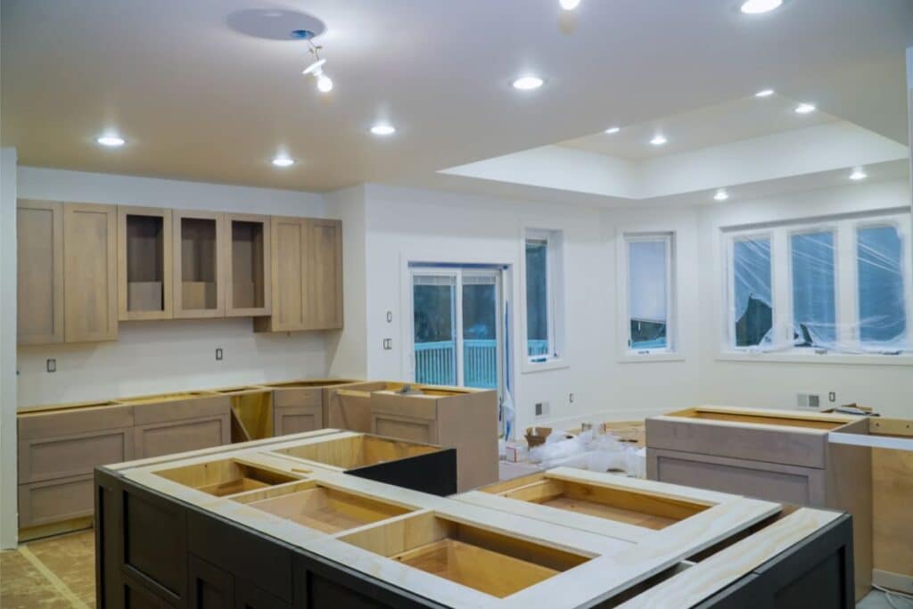 Modern kitchen renovation project under construction with unfinished wooden cupboards, an empty kitchen island, and recessed lighting. Windows and sliding doors provide natural light, whilst some surfaces are covered for protection.