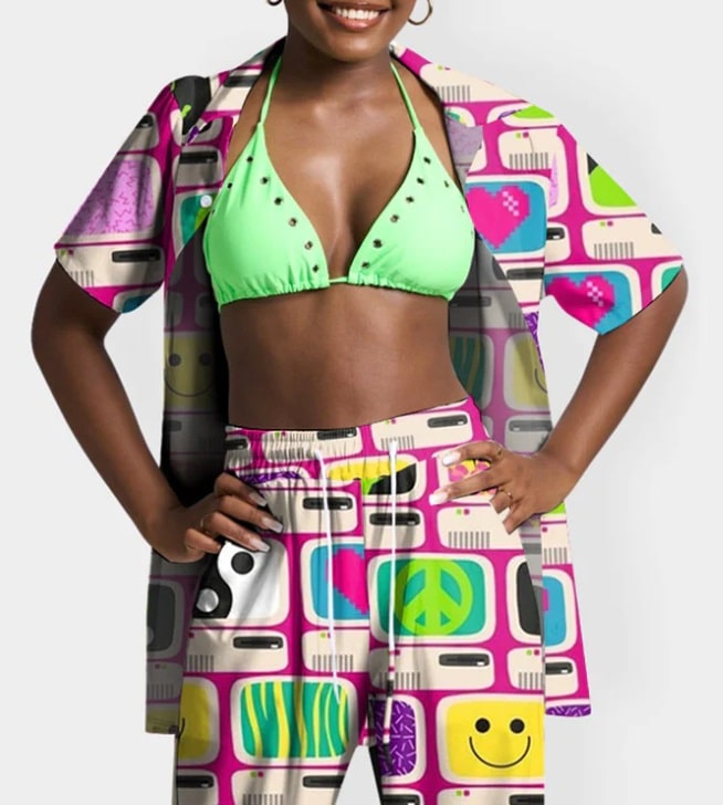 A person smiles whilst wearing a green bikini top and a matching open shirt and trousers set from Cherry Kitten, adorned with colourful, retro patterns featuring peace signs, smiley faces, and hearts. The background is plain white.