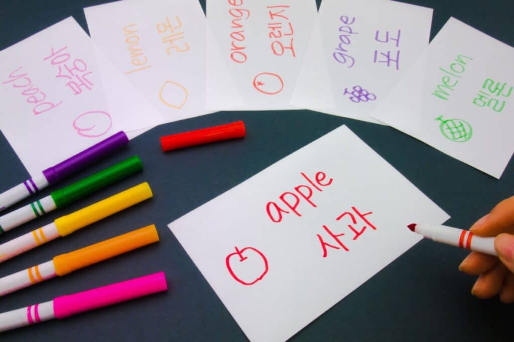 Bilingual flashcards show fruit names in English and Korean, with colourful drawings. Cards read "apple," "peach," "lemon," "orange," "grape," and "melon." Coloured markers and a hand holding a red marker are nearby.