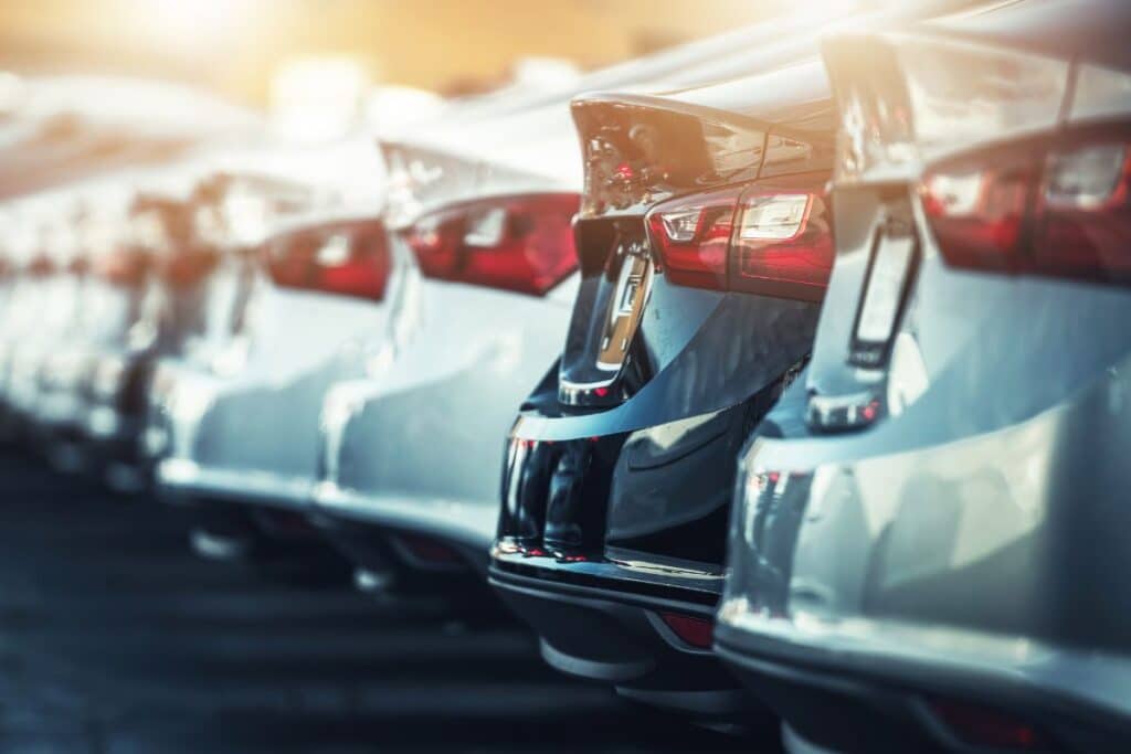 Row of parked silver cars seen from the rear, with sunlight reflecting off each car’s glossy surface. The cars are lined up closely, emphasising their taillights and rear bumpers in a bright, unified display.