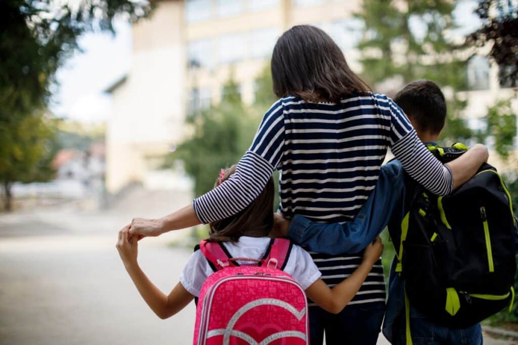 A woman with her arm round two children, one girl with a pink rucksack and one boy with a green rucksack, enjoys a stress-free school run as they walk together outdoors towards a building, their backs to the camera.