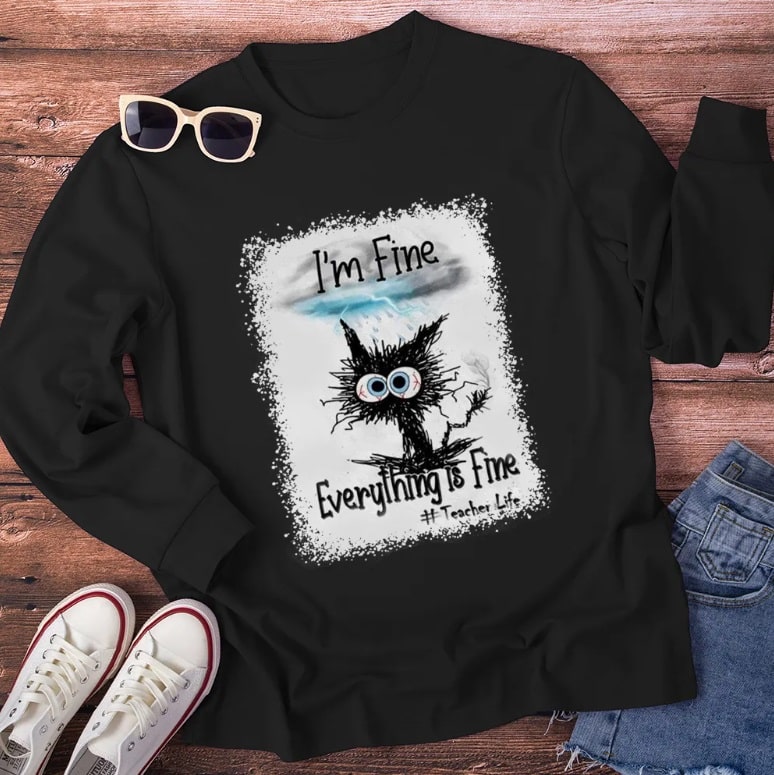 A black sweatshirt with a cartoon of a frazzled black cat and text reading "I'm Fine, Everything Is Fine #TeacherLife." Show off your style by pairing it with white trainers, denim shorts, and sunglasses on a wooden surface.