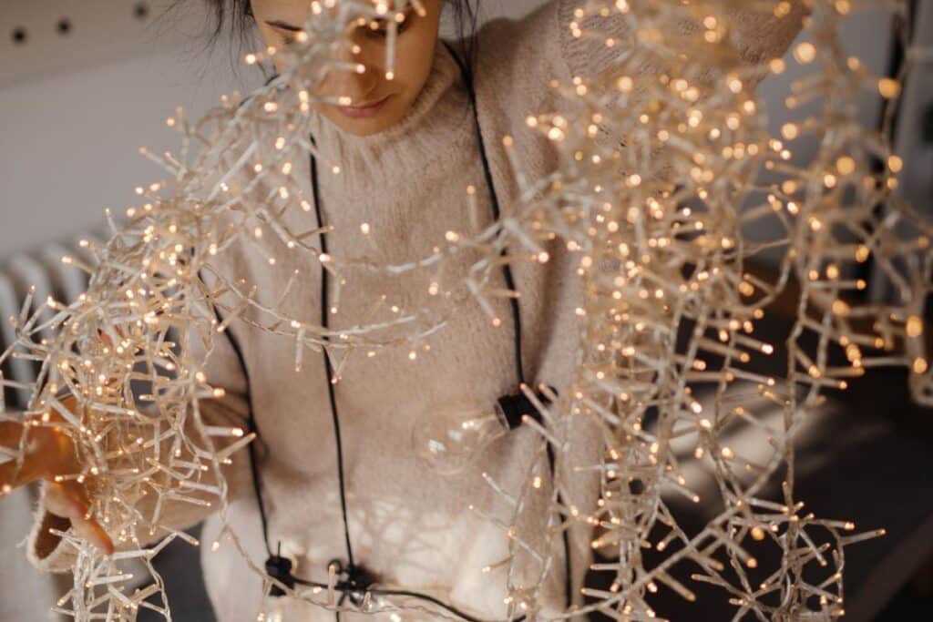 A person wearing a light-coloured jumper untangles a large string of lit Christmas lights, holding them up in front of their face in a softly lit indoor setting.