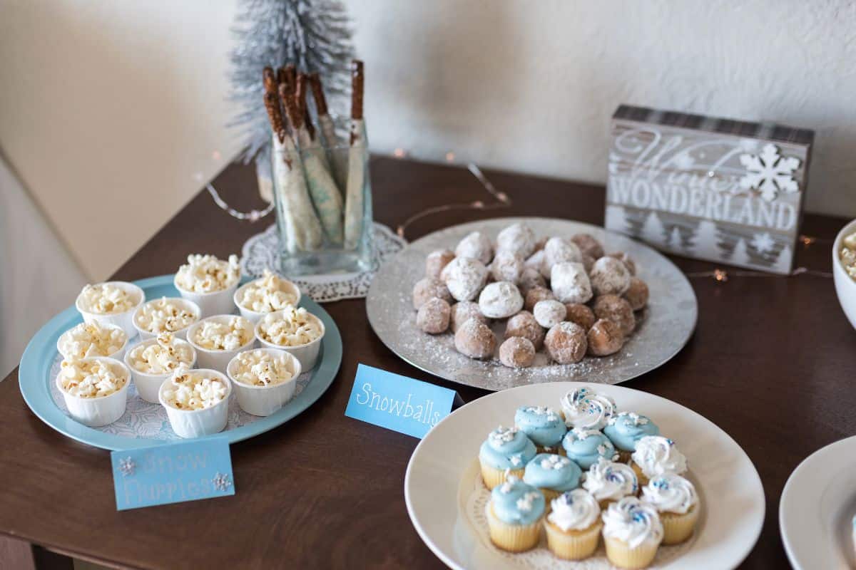 A winter-themed dessert table for a birthday party in winter with plates of popcorn, icing sugar doughnut holes, blue and white iced cupcakes, biscuit sticks in a glass, and a “Winter Wonderland” sign in the background.